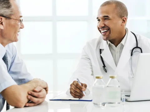 A senior man visits his male doctor for his annual screening. Both are seated at a table.